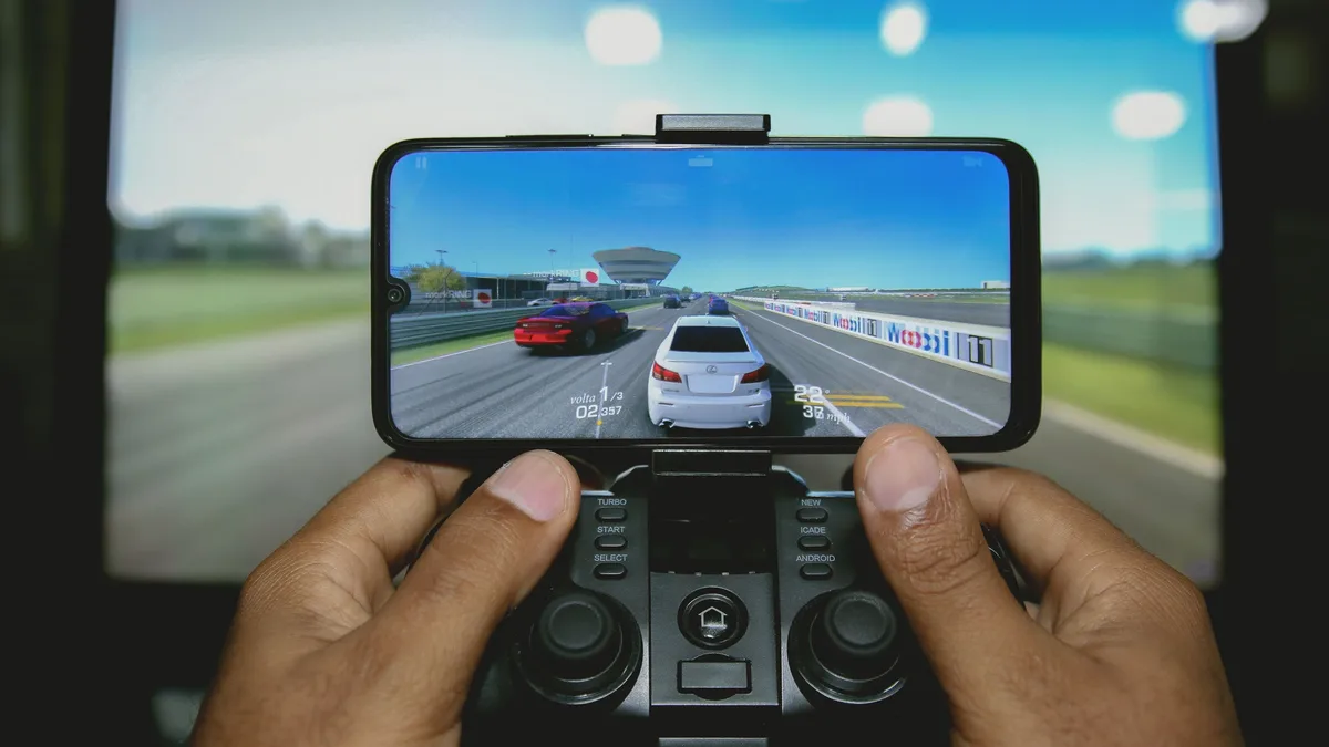 A person is holding a smartphone with a video game on the screen, and their thumbs are on a controller attached to the phone, with a large screen displaying a racing game in the background.