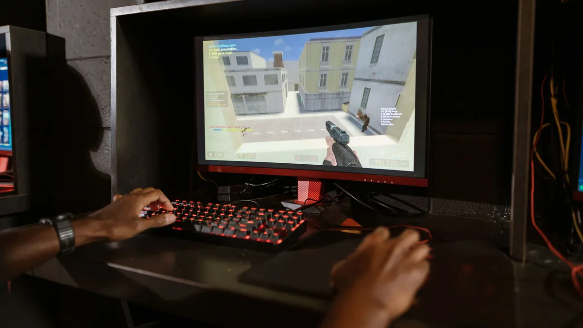 A person playing a video game on a computer monitor, with the monitor displaying a first-person shooter game featuring a gun-wielding character in a building.